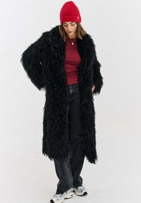 Černý huňatý kabát s dlouhými rukávy, nošený přes vínově červený top a černé džíny. Osoba má na hlavě červenou čepici a bílé tenisky.