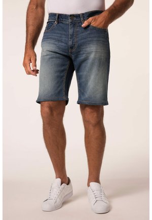 Mand iført blå denimshorts og hvide sneakers står med den ene hånd i lommen mod en ensfarvet baggrund.