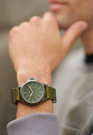 Grøn analog ur med et rundt, sølvfarvet metalhus, store hvide tal og et olivengrønt stofrem, båret på et håndled med en grå ærme.