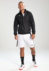 Veste zippée légère noire, short blanc avec des bandes latérales noires, associé à des chaussettes noires et des chaussures de football blanches et rouges. Logo Puma visible.