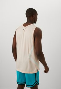 Leicht pfirsichfarbener Mesh-Trägershirt mit kontrastierenden hellblauen Akzenten. Enthält ein kleines schwarzes Logo. Getragen mit türkisfarbenen Shorts mit schwarzen Streifen.