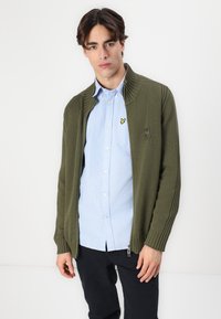 Olivengrønn strikket cardigan med ribbede kanter, full glidelås og en liten logo-patch. Brukt over en lys blå skjorte med knapper.