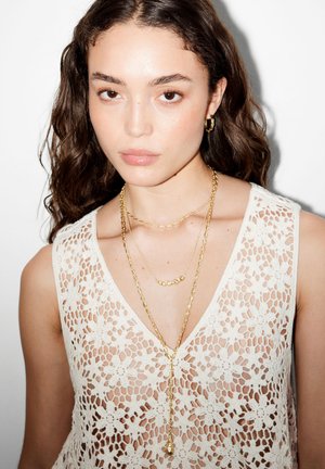 Junge Frau mit lockigem braunem Haar, die ein weißes, mit Blumen versehenes Spitzentop und geschichtete goldene Halsketten sowie Creolen trägt, vor einem weißen Hintergrund.