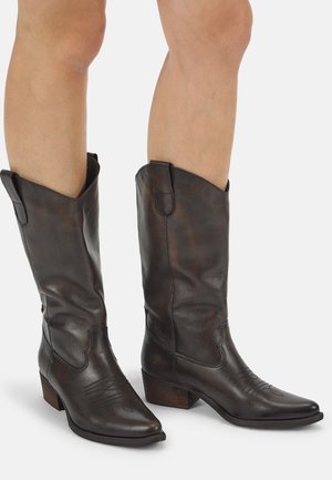 Beine mit dunkelbraunen Leder-Mid-Calf-Westernstiefeln mit niedrigen Holzabsätzen und gestickten dekorativen Mustern.