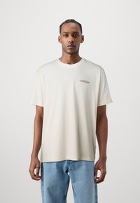 Világos bézs pamut póló kerek nyakkivágással, rövid ujjakkal, és egy kis piros "GUESS JEANS" logóval a bal mellkason. Kék farmernadrággal viselve.