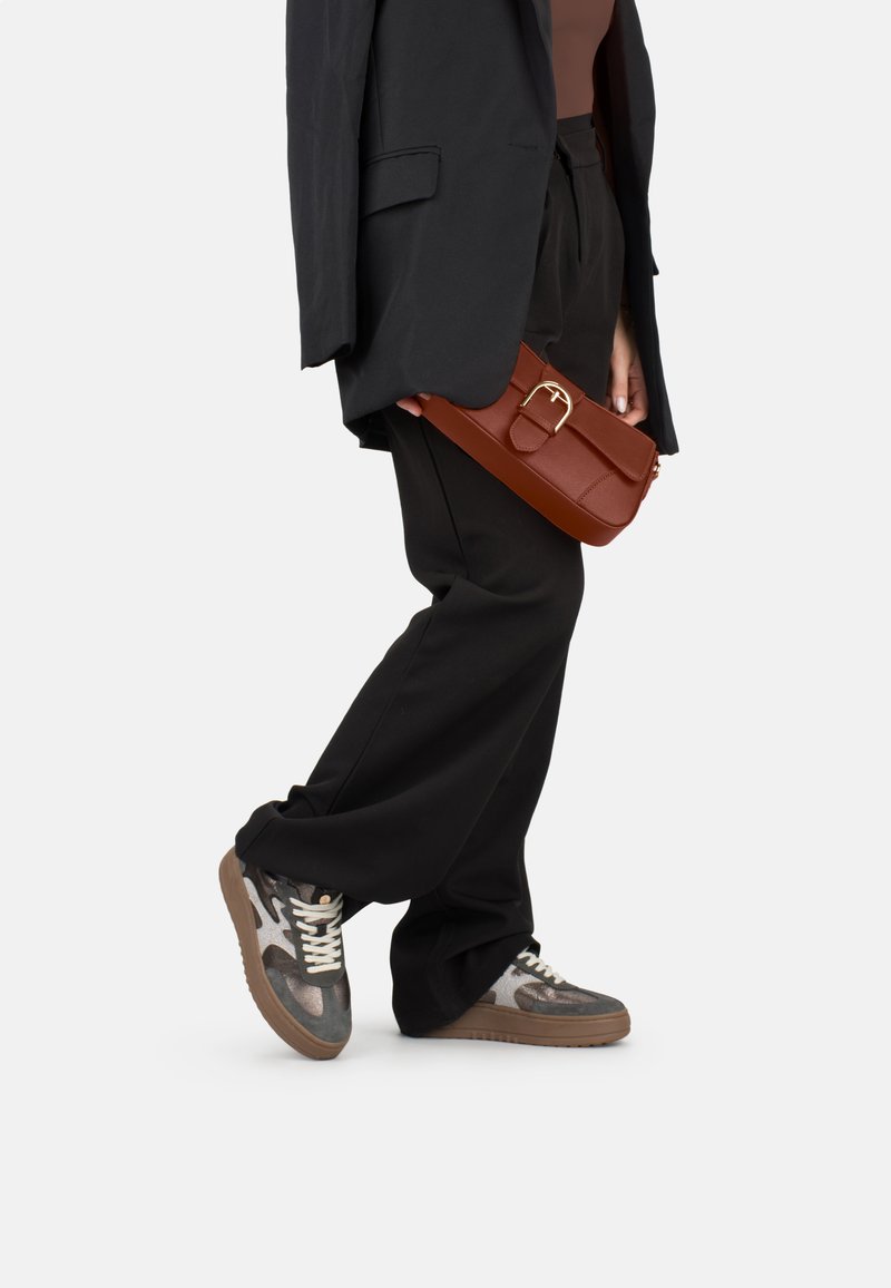 Braune Kunstleder-Crossbody-Tasche mit rechteckiger Form, flachem Deckel und goldfarbigem Schnallendetail. Kombiniert mit lockeren schwarzen Hosen und gemusterten Turnschuhen.