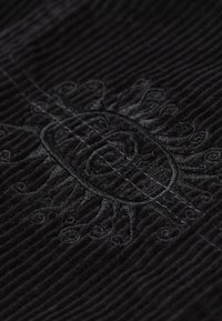 Schwarzer Cordstoff mit kunstvoller schwarzer Stickerei eines Sonnenmotivs, verziert mit Wirbeln und musikalischen Noten entlang strukturierter Linien.