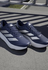 Scarpe da corsa navy con tomaia in rete. Presentano tre strisce bianche su ciascun lato, superficie testurizzata e un design della suola moderno e arrotondato.