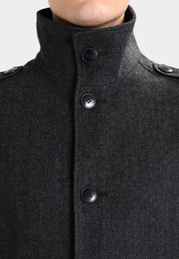 Abrigo de espiga gris oscuro con un alto cuello, que presenta botones negros y hombreras. Fabricado con una tela texturizada similar a la lana.