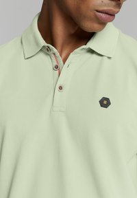 Polo shirt in lichtgroene katoen, met een kraag, drie knopen met roze accenten en een zwart hexagonaal logo op de borst.