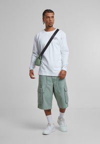 Weißes Langarmhemd, hellgrüne Cargo-Shorts, kombiniert mit weißen Sneakers und einer kleinen grünen Tasche. Die Textur ist glatt, das Design ist lässig.