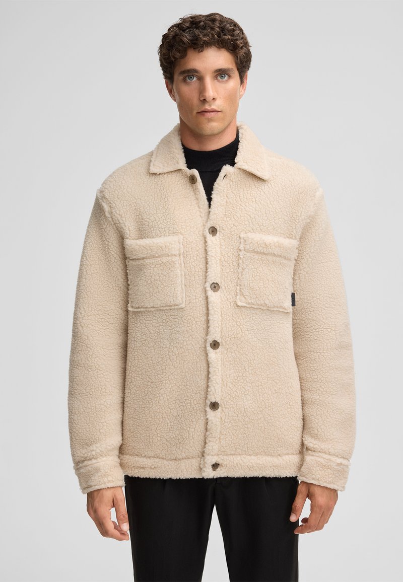 Beige fleece jacket with a textured surface, featuring a collar, front buttons, and two chest pockets. The sleeves are cuffed.