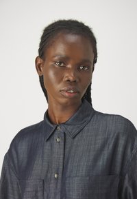 Camicia in denim blu scuro con colletto, due tasche sul petto e chiusura con bottoni. Il tessuto presenta una sottile texture e una leggera lucentezza.
