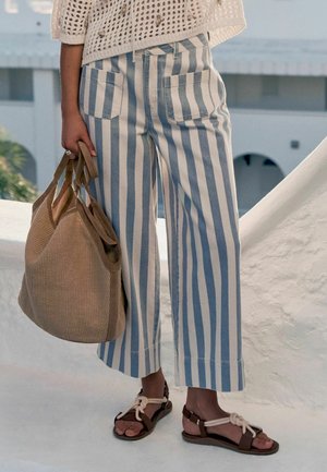 Frau trägt blau-weiß vertikal gestreifte Hose, hält eine beige geflochtene Tragetasche und trägt braune Sandalen mit weißen Seilriemen.