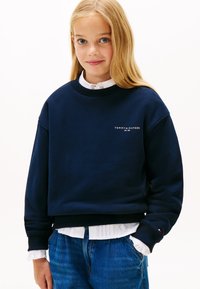Felpa blu navy in cotone con colletto e polsini a costine; presenta il logo bianco "TOMMY HILFIGER" sul davanti, indossata con una camicia bianca con colletto.