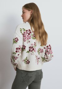 Suéter de punto con base blanca, con patrones florales en burdeos y verde. Dobladillo y puños acanalados. Se lleva con pantalones grises.