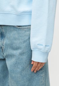 Χέρι που ακουμπάει στο πλάι, φορώντας ανοιχτό μπλε φούτερ με ριμπ μανσέτα και ανοιχτό μπλε τζιν με ορατό ραφή τσέπης.