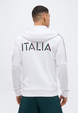 Mann, der mit dem Rücken zur Kamera steht, trägt einen weißen Kapuzenpullover mit Kapuze, auf dessen Rückseite "ITALIA" in roten, grünen und schwarzen Buchstaben aufgedruckt ist.