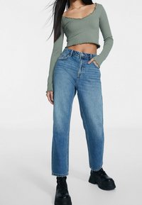 Top verde de manga larga con texto acanalado y bordes festoneados, combinado con jeans de pierna recta de tiro alto en azul claro y botas gruesas negras.