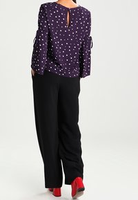Blusa a pois viola con schiena a chiave, abbinata a pantaloni neri a gamba larga. Stivali alla caviglia rossi mettono in risalto l'outfit.