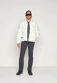 Άτομο που φορά μαύρα γυαλιά ηλίου, λευκή πούφκα, μαύρο μπλουζάκι, σκούρο παντελόνι και μαύρες μπότες, στέκεται μπροστά σε απλό ανοιχτό φόντο.