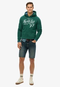Haut à capuche vert avec imprimé graphique blanc, associé à un short en jean, des baskets crème et des chaussettes multicolores. Style décontracté et coupe ample.