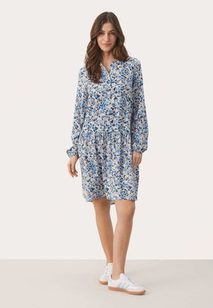 Bloemenjurk van lichte stof met blauw-witte patronen. Voorzien van een kraag, een geplooide taille en lange mouwen. Gecombineerd met witte sneakers.