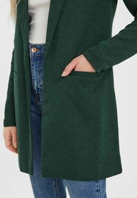 Dunkelgrüner langer Cardigan aus weichem Stoff mit offenem Vorderteil, Seitentaschen und langen Ärmeln, getragen über einem weißen Oberteil und blauen Jeans.