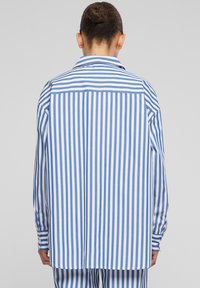 Camicia a righe blu e bianca, con maniche lunghe, taglio dritto e polsini con bottoni. La vista posteriore mostra un dettaglio della giogaia a contrasto.