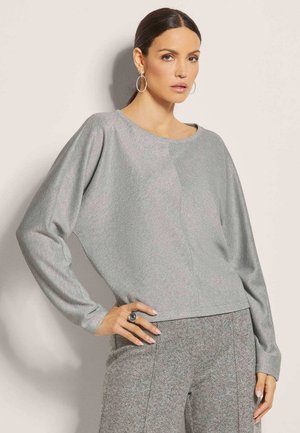 Femme aux cheveux foncés attachés porte des boucles d'oreilles créoles argentées, un haut à manches longues gris clair et un pantalon gris texturé, posant avec la main sur la hanche.