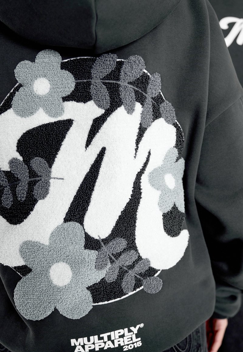 Černá mikina s kapucí s jemným bílým a šedým květinovým vzorem kolem velkých stylizovaných písmen "Mc" a textem "Multiply Apparel 2015" pod nimi.