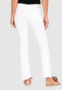 White flared jeans with a raw hem and two back pockets, worn with light brown sandals featuring a flower accent and ankle ties.