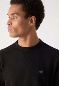 Sweatshirt preto com gola redonda, colarinho texturizado canelado e um pequeno logotipo no peito. Tecido suave, design casual.