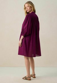 Robe couleur prune avec une coupe ample, des manches bouffantes, des détails de poignet à boutons et une longueur au genou. Portée avec des sandales à imprimé léopard.