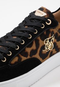 Baskets en imprimé léopard noir et marron avec des accents dorés, présentant des lacets noirs plats, un matériau en suède et un design texturé. Semelle blanche.