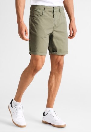 Mężczyzna ubrany w oliwkowozielone szorty z mankietami, białe skarpetki sięgające za kostkę oraz białe trampki z gumowymi podeszwami, stojący na jednolitym białym tle.