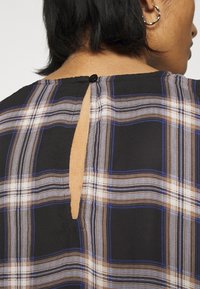 Personne portant un haut à carreaux noirs, marron et bleu avec une petite fermeture à bouton et une ouverture en forme de trou de serrure dans le dos au niveau de la nuque, montrant des cheveux noirs courts.