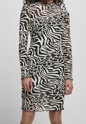 Kvinde iført en knælængde kjole med lange ærmer med sort-hvidt abstrakt zebra print, der står mod en ensfarvet baggrund.