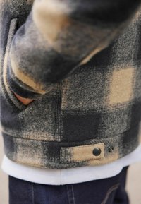 Uldjakke med et sort og beige ternet mønster, har en sidelomme og en trykknapdetalje ved taljen. Blød, tekstureret overflade.