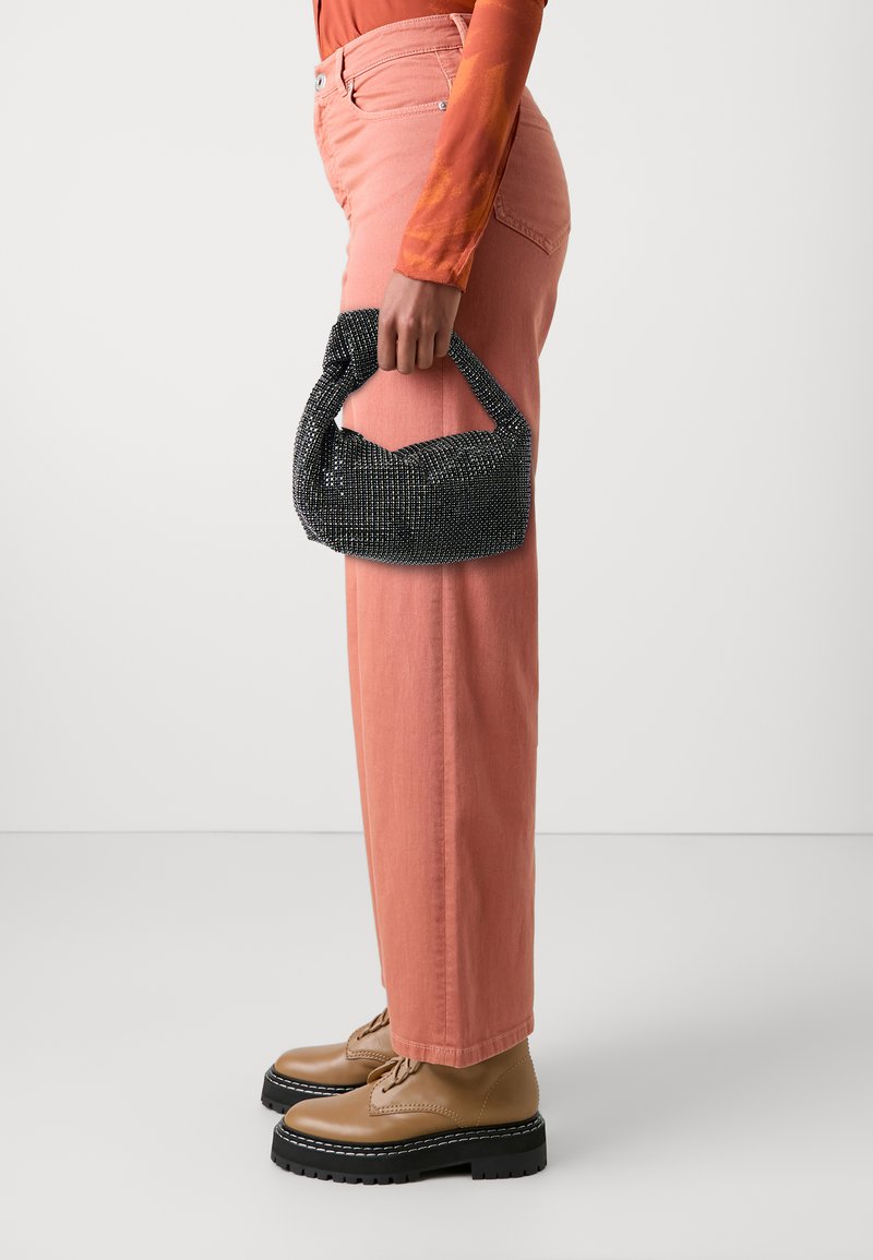 Black beaded handbag with a knotted handle, held in a hand. The person wears light pink trousers and tan platform shoes.