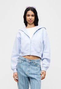 Νέα γυναίκα που φοράει ανοιχτό μπλε cropped φούτερ με φερμουάρ, λευκή μπλούζα, ριγέ ανοιχτό μπλε τζιν ψηλόμεσο και γυαλιά ηλίου στο κεφάλι.