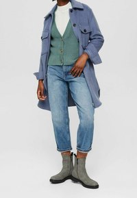 Jasnoniebieski wełniany płaszcz z dużymi kieszeniami, zielony dziergany sweter z guzikami, niebieskie dżinsy i szare botki za kostkę z bocznym zamkiem.