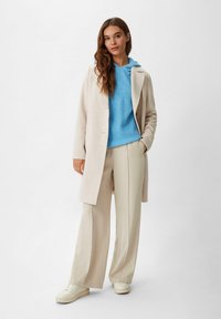 Helle beige Jacke über einem blauen Strickhoodie, kombiniert mit beigen, weit geschnittenen Hosen. Glatte Textur, simples Design und lässiges Schuhwerk.