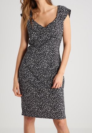 Kobieta ubrana w czarno-białą, nakrapianą, bezrękawną sukienkę do kolan z dekoltem w serek i falbanami na ramionach.