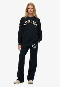 Sort sweatshirt med "SUPERDRY" i cremefarvet tekst, parret med sorte sweatpants der har "DEPT 86" i creme. Blød bomuld, afslappet pasform.