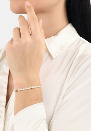Pulsera de oro y plata con cuentas redondas y acentos en forma de corazón, llevada en una mano, combinada con una camisa blanca de cuello. Textura suave.