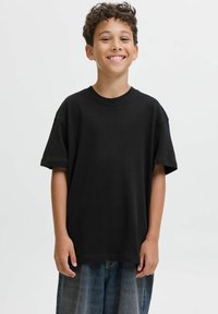 Un garçon souriant avec des cheveux bouclés portant un t-shirt noir ample et un short en denim bleu se tenant devant un fond clair.