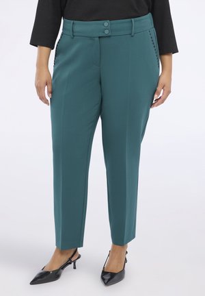 Pantalons cintrés teal avec deux boutons sur le devant, des détails en clous sur les côtés et une texture lisse, associés à des chaussures noires à bout pointu.