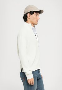 Mlađi muškarac nosi bež kapu, bijeli duksericu s patentnim zatvaračem preko prugaste košulje i plave traperice, okrenut desno prema jednostavnom bijelom pozadinskom.