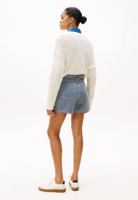 Suéter blanco de punto trenzado sobre una camisa de mezclilla, combinado con pantalones cortos a rayas azules y blancas. Modelo lleva zapatillas blancas con suela marrón.